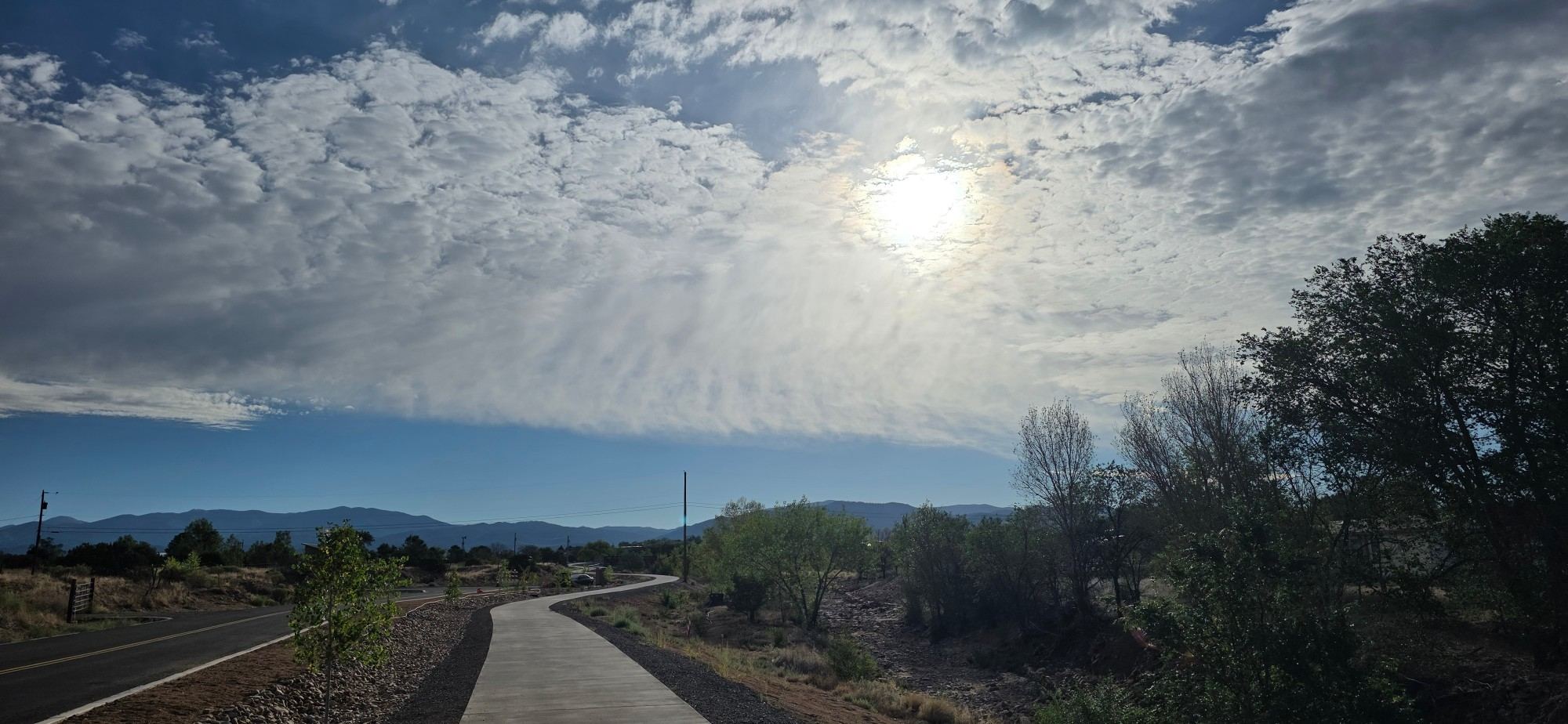 The paved Santa Fe River Trail curving beside a road under a wide, cloud-filled sky, with trees along one side and mountains on the horizon